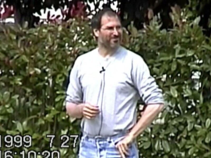Steve Jobs speaks at an Apple employee gathering in 1999, with a screen displaying a product behind him.